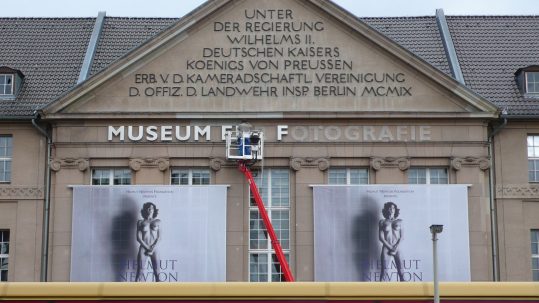 Museum für Fotografie, Berlin, Teil der Fassade, Hubbühne bei der Arbeit und große Plakate mit einem Fotomotiv von Helmut Newton, davor das Dach einer S-Bahn
